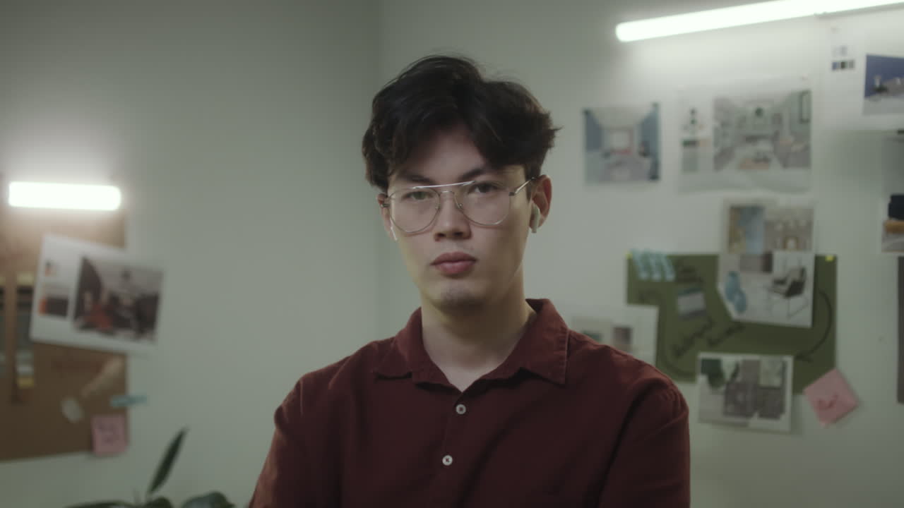 A young Asian man wearing glasses and earbuds in an office setting
