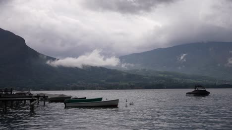 Barcos-Flotando-En-El-Lago-Annecy-En-Los-Alpes-Franceses-Con-Montañas-Nubladas-Al-Fondo,-Plano-Amplio