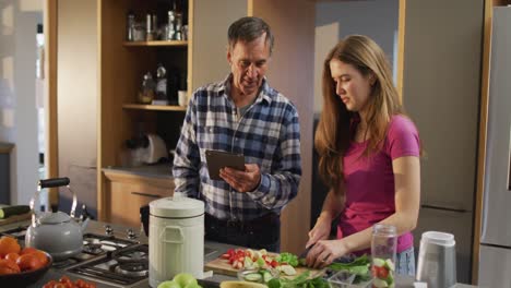 padre caucásico mayor sonriente con su hija adolescente preparando una bebida saludable usando una tableta en la cocina