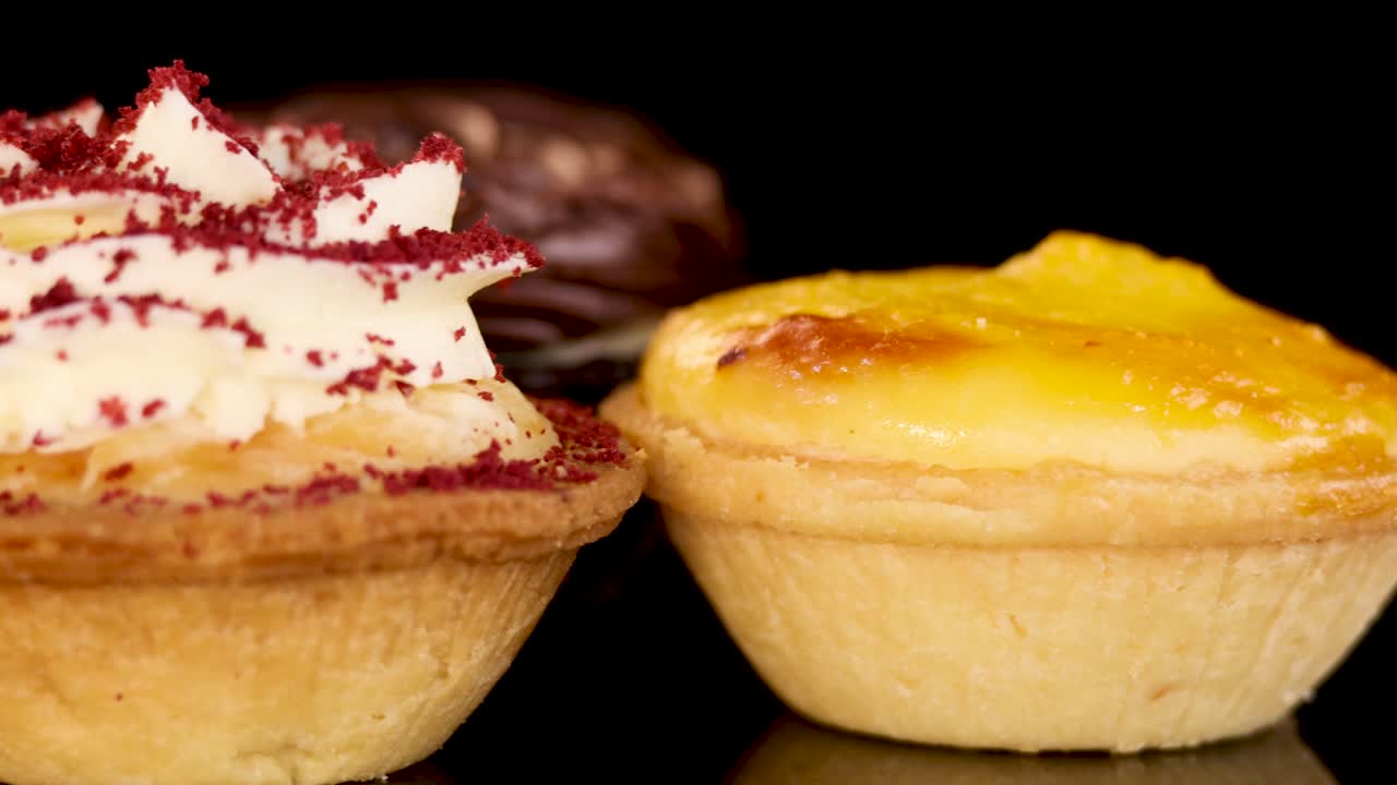 Close-up rotating view of assorted mini baked desserts, including chocolate cream tarts, egg tarts, and red velvet cupcakes, on a reflective black surface with soft studio lighting