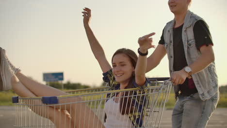 Cupé-Joven-Y-Elegante-Divirtiéndose-Montando-Con-Un-Carrito-De-Compras-En-El-Estacionamiento-Al-Aire-Libre-Cerca-Del-Supermercado