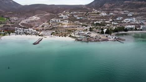 Toma-Panorámica-Aérea-Sobre-La-Hermosa-Costa-De-Playa-El-Caymancito-Cerca-De-La-Paz-Baja-California-Sur-México-Con-Vista-Al-Paisaje-Seco-Y-Al-Edificio-Del-Hotel-Con-Mar-Turquesa