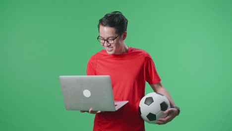 hombre asiático con una pelota viendo un partido de fútbol en una computadora portátil en el estudio de pantalla verde