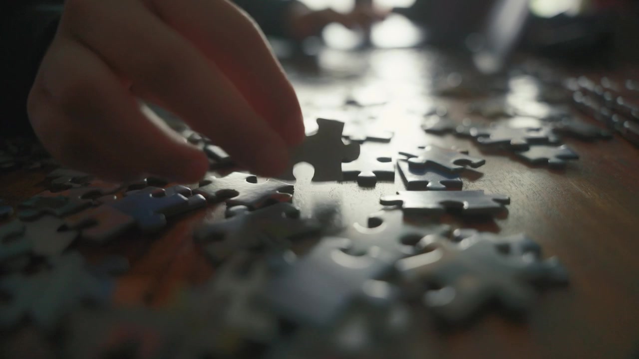 Un primo piano fisso a livello del tavolo mostra una mano che seleziona pezzi di puzzle tra forme sparse. La profondità di campo ridotta e la retroilluminazione soffusa creano una scena intima ideale per contenuti dedicati agli hobby e alla risoluzione di problemi.