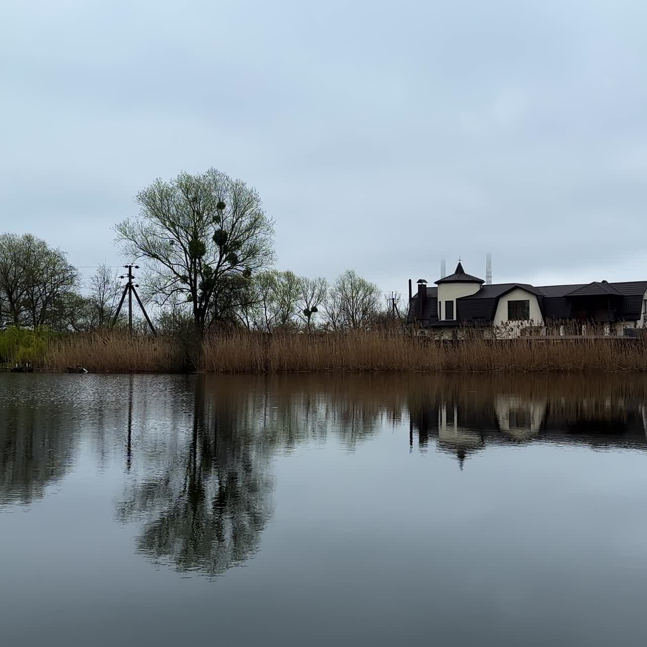 Chilly day on the calm river. Beautiful house on the bank overgrown with dry grass and bare trees in spring