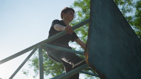 a young boy sits on an outdoor blue iron structure, lost in thought, with both hands gripping the framework, sunlight filters through the lush green trees behind him