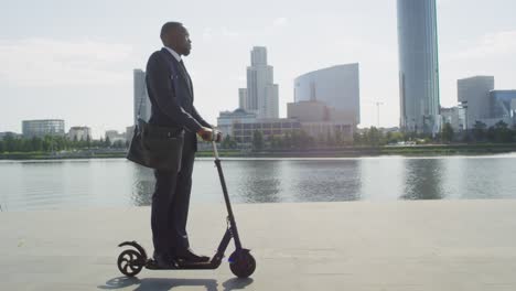 young serious businessman on electric scooter by riverside against tall modern buildings