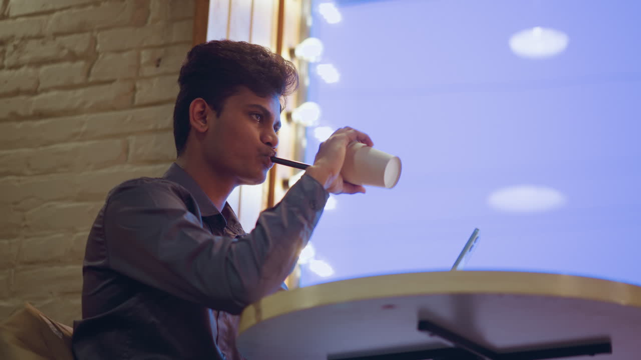 Giovane uomo seduto in un caffè moderno che sorseggia l'ultima goccia da una tazza di carta mentre guarda lo smartphone, circondato da luci a specchio soffuse vicino a una grande finestra, mostrando una postura rilassata e un'atmosfera urbana tranquilla.