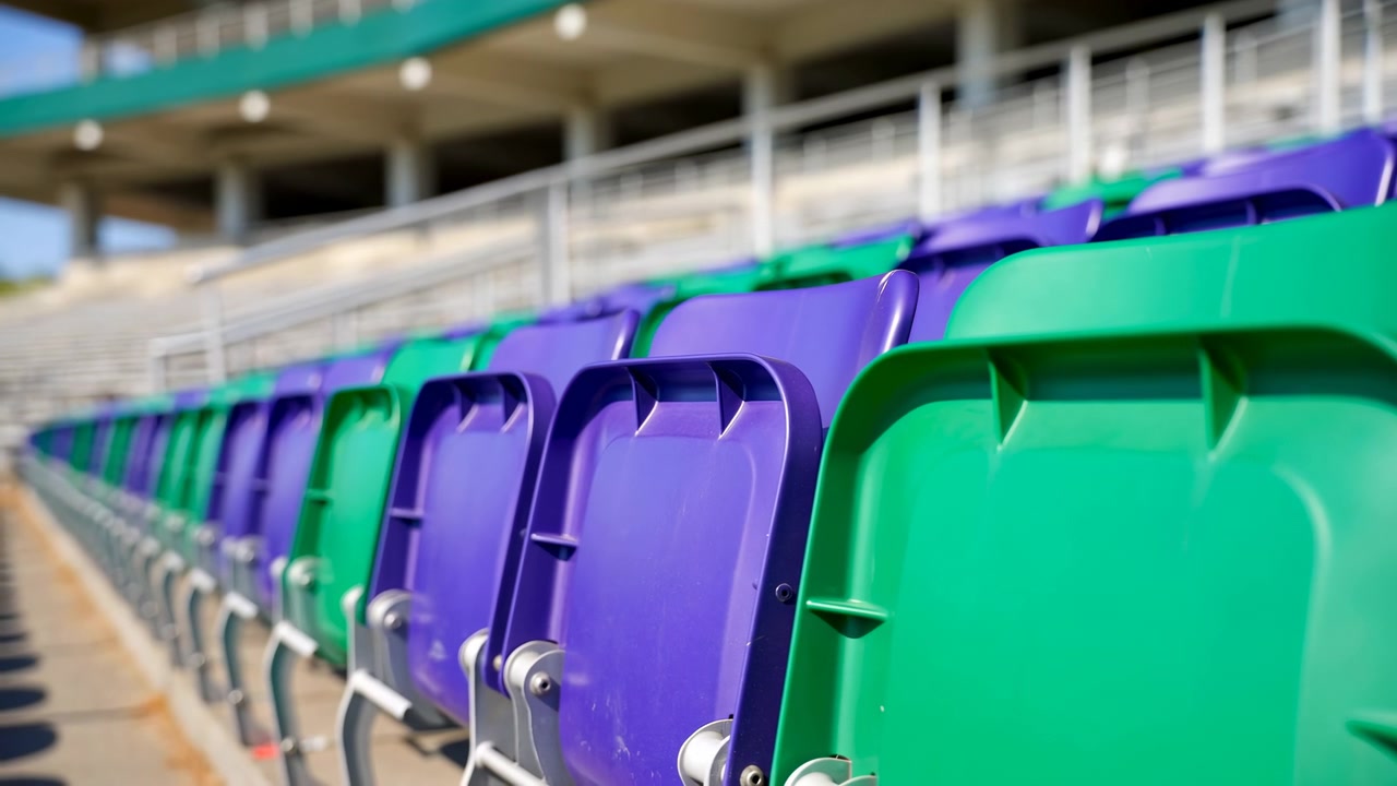 Nahaufnahme in Bewegung von abwechselnd violetten und grünen geformten Plastikstadionsitzen, die diagonal über leere Tribünen im Freien verlaufen, mit scharfem Fokus im Vordergrund auf Rückenlehnen, Oberkanten und Metallscharniere.
