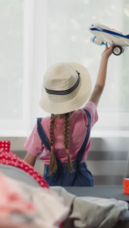 little girl plays with toy plane standing by window while mother folds clothes in stack on sofa in living room. kid dreams about family vacation