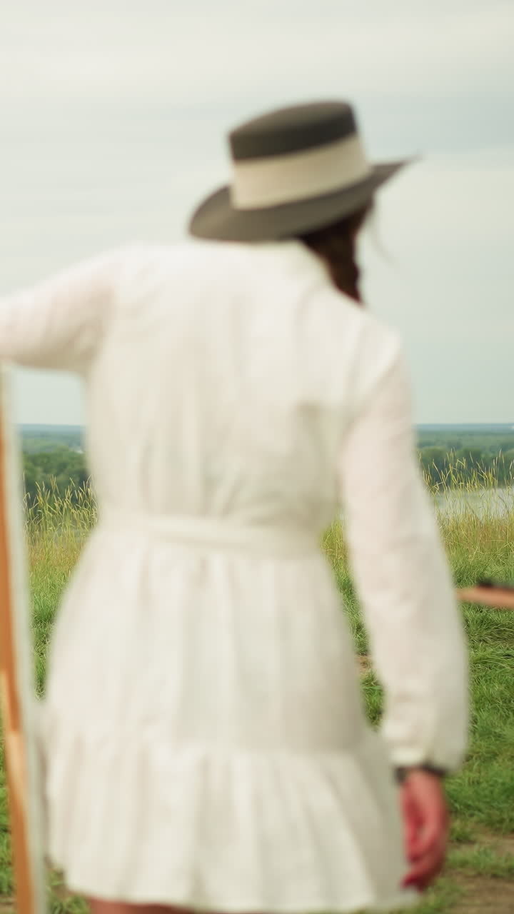 vista trasera de una pareja caminando por un campo cubierto de hierba. la mujer, con una túnica blanca, lleva un lienzo mientras que el hombre, con una camisa a cuadros y vaqueros, sostiene un bloc de dibujo y un caballete
