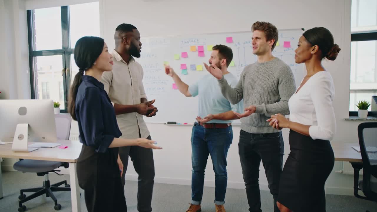 Leading man raising hands, team of five in office wear planning around whiteboard with sticky notes