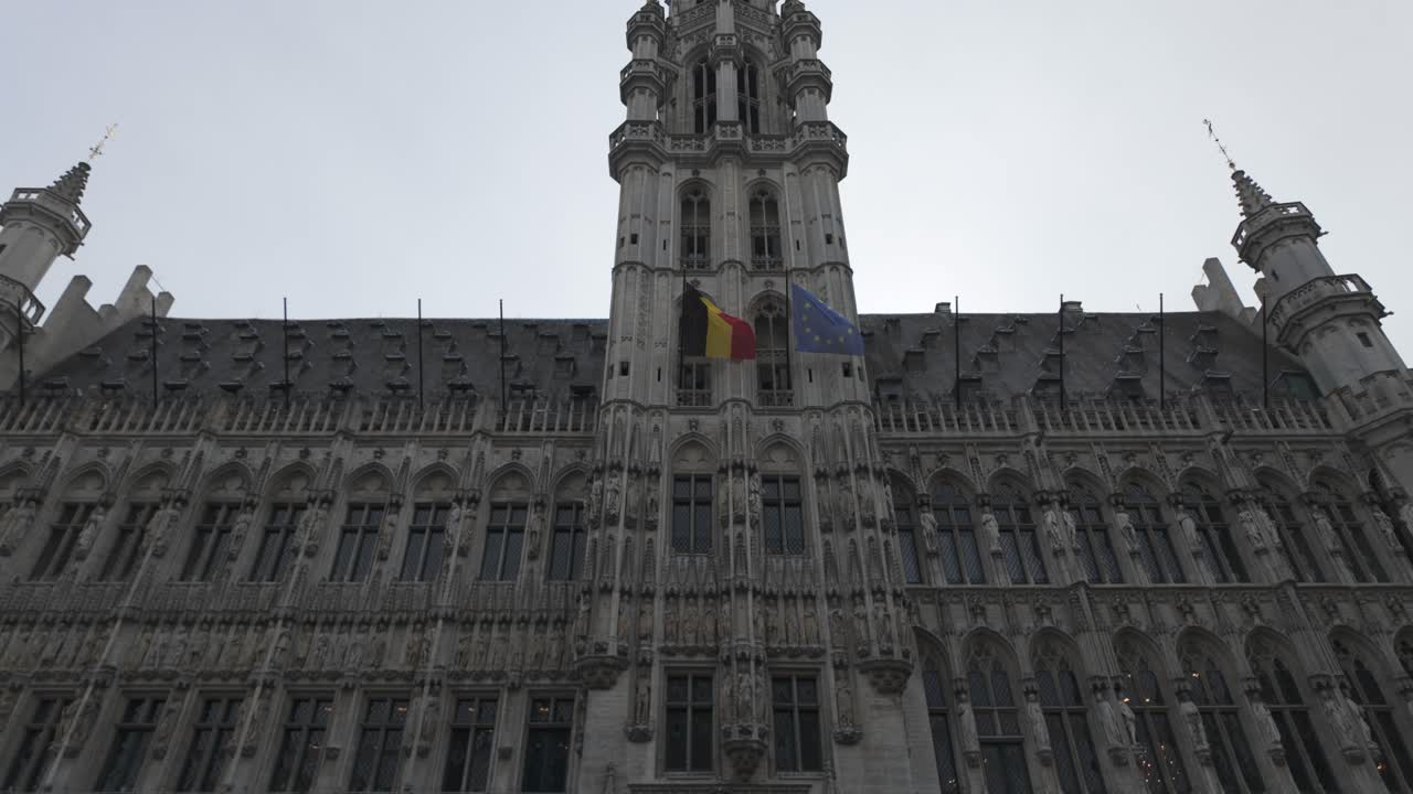 The facade of Brussels Town Hall with Belgian and European Union flags waving on a cloudy day. pan up shot
