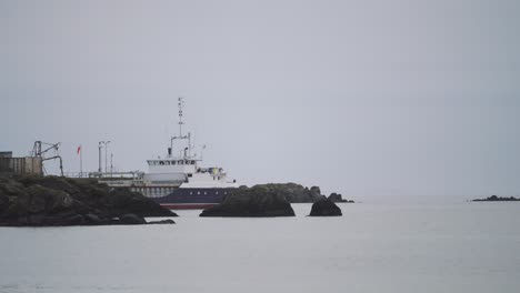 Boat-Sitting-In-Grey-Water-and-Sky-Surrounded-By-Rocks