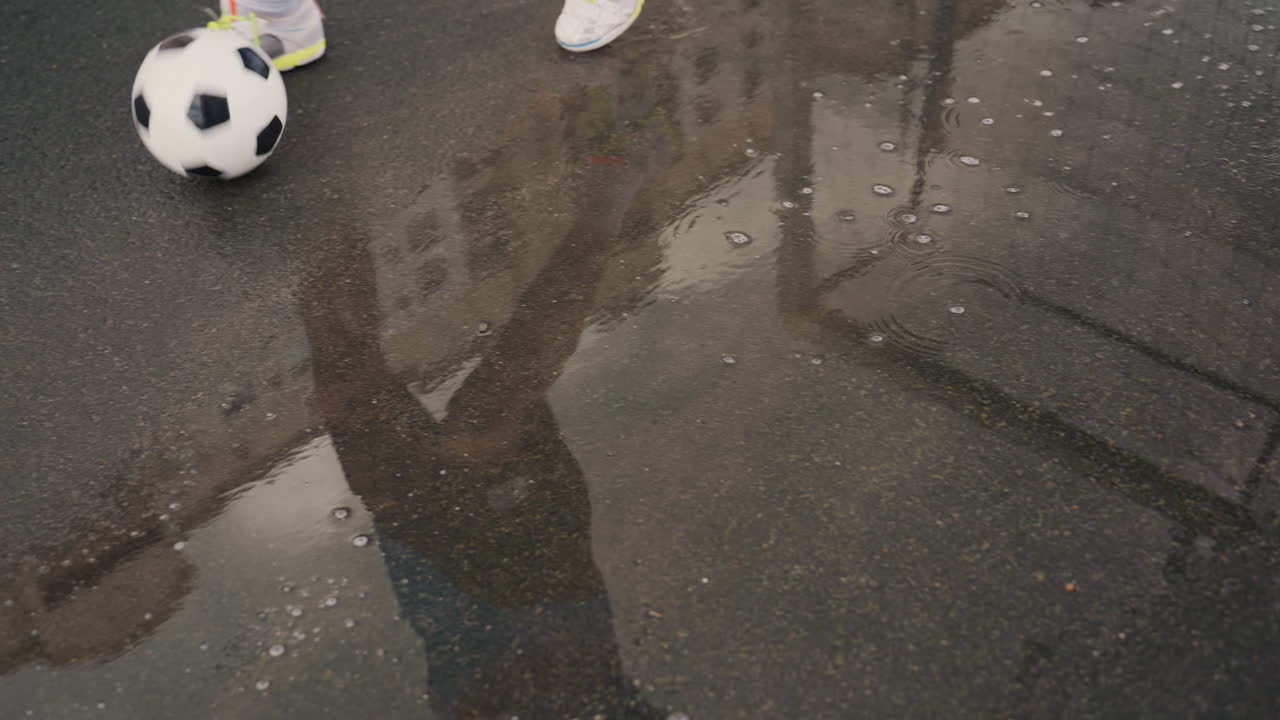 Eine nasse Asphaltpfütze, in der ein Fußballspieler zu sehen ist, der in einem städtischen Regen den Ball dribbelt, Turnschuhe spritzen, Stadtgebäude spiegeln sich auf der Oberfläche, trübes, bedecktes Licht, Nahaufnahme von Beinen und Ball auf der Straße