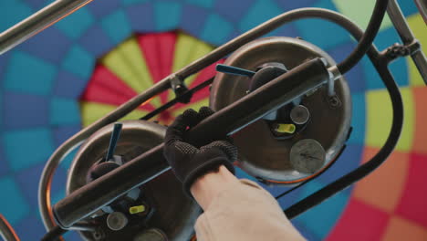 close up of gloved hand gripping control bar beneath metal gas burner system with colorful hot air balloon canopy above showing vibrant fabric pattern mechanical parts