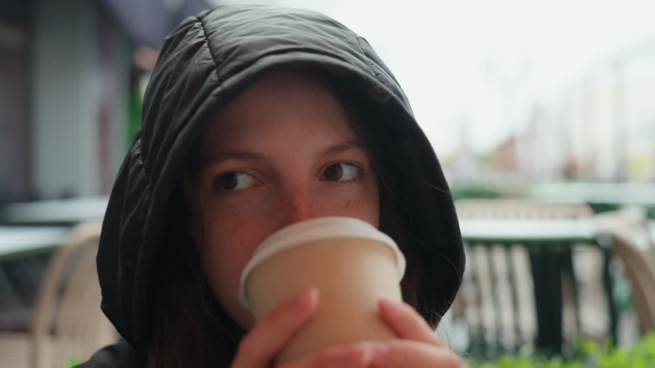 フードを被った女性が屋外のテラスカフェでコーヒーを飲み、朝の光の中で物思いにふける様子。そばかすがあり、温かいカップを手に持ち、小雨から身を守るニットのフーディーを着ている。映画のような浅い被写界深度で、静かな都会の雰囲気が伝わる、自然な一枚。