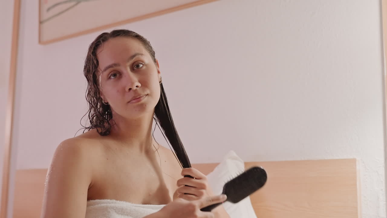 Donna con capelli bagnati, asciugamano, letto d'hotel, pettinatura delicata dopo la doccia, trecce sciolte che cadono, luce calda e soffusa, arredamento minimalista della stanza, atmosfera mattutina rilassata, goccioline d'acqua sulle ciocche, cura di sé informale.