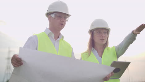 Coworking-engineers-with-tablets-on-solar-plant.-Adult-men-and-women-in-hardhats-using-tablets-while-standing-outdoors-on-transformer-platform.-Transportation-of-clean-energy.-Wind-energy-delivery