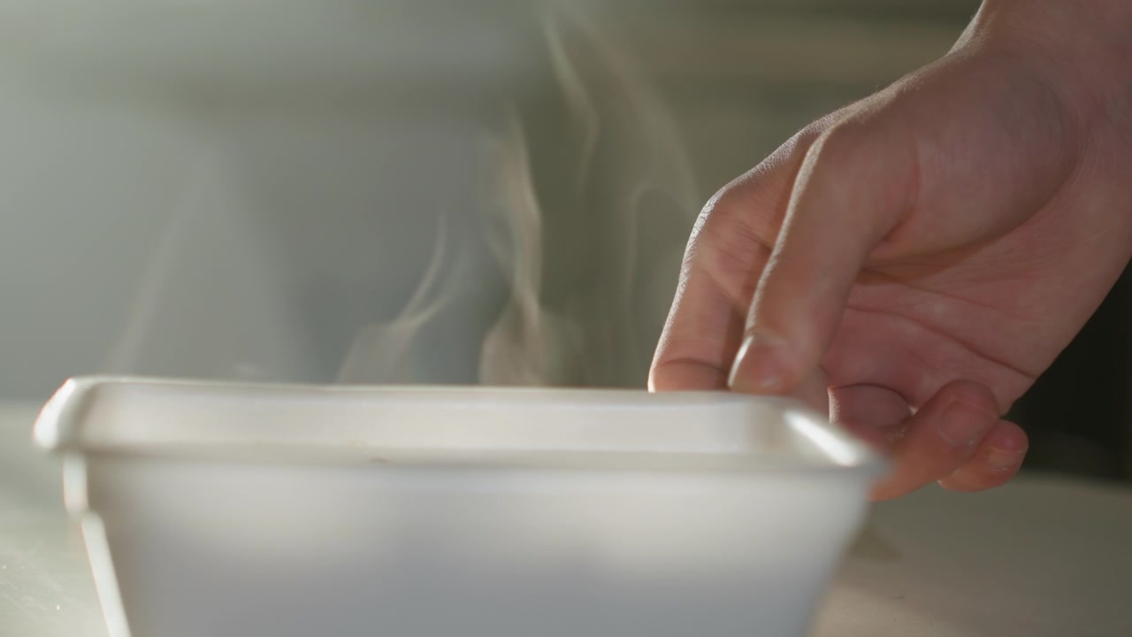 Hånd som tester varm, dampende beholder på kjøkkenbenken, nærbilde av fingre som svever over plastboks, synlig damp som stiger, mykt morgenlys, forsiktig bevegelse som formidler varme og trygghet