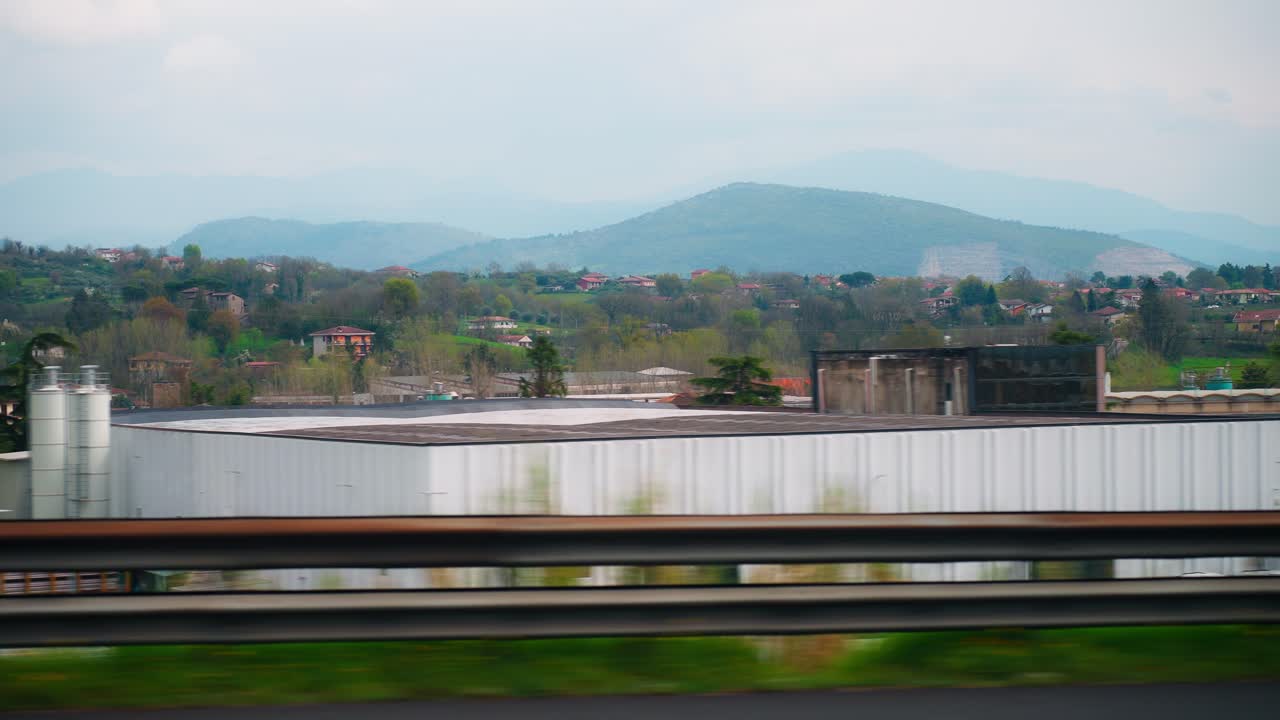 View from the road between Naples and Pompeii, with guardrail, scattered buildings, industrial tanks, and hazy hills passing by under an overcast sky