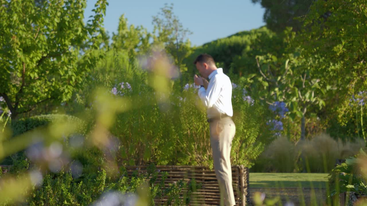 베이지 색 바지, 흰색 셔츠, 짧은 갈색 머리를 가진 젊은 백인 남자가 녹지로 가득한 햇볕이 잘 드는 공원을 걷고 작은 꽃 냄새를 맡을 것입니다
