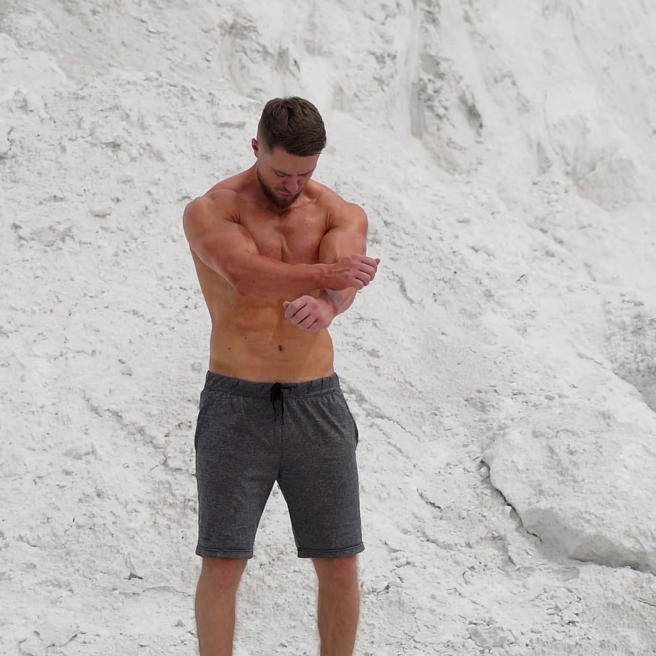 Bearded man with muscular body poses on white mountain background. Strong bodybuilder outdoors in summer.