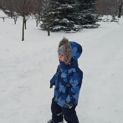 caucasian baby wearing blue coat with fur hood walks outdoor, speaks and points at something. walk in the park in winter.