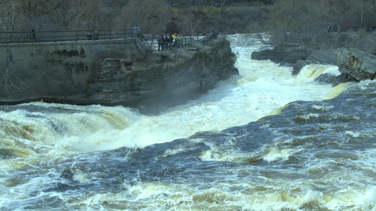 Swollen rocky river with high cliffs. Water bubble away in slow motion as a group of people observe from a lookout above