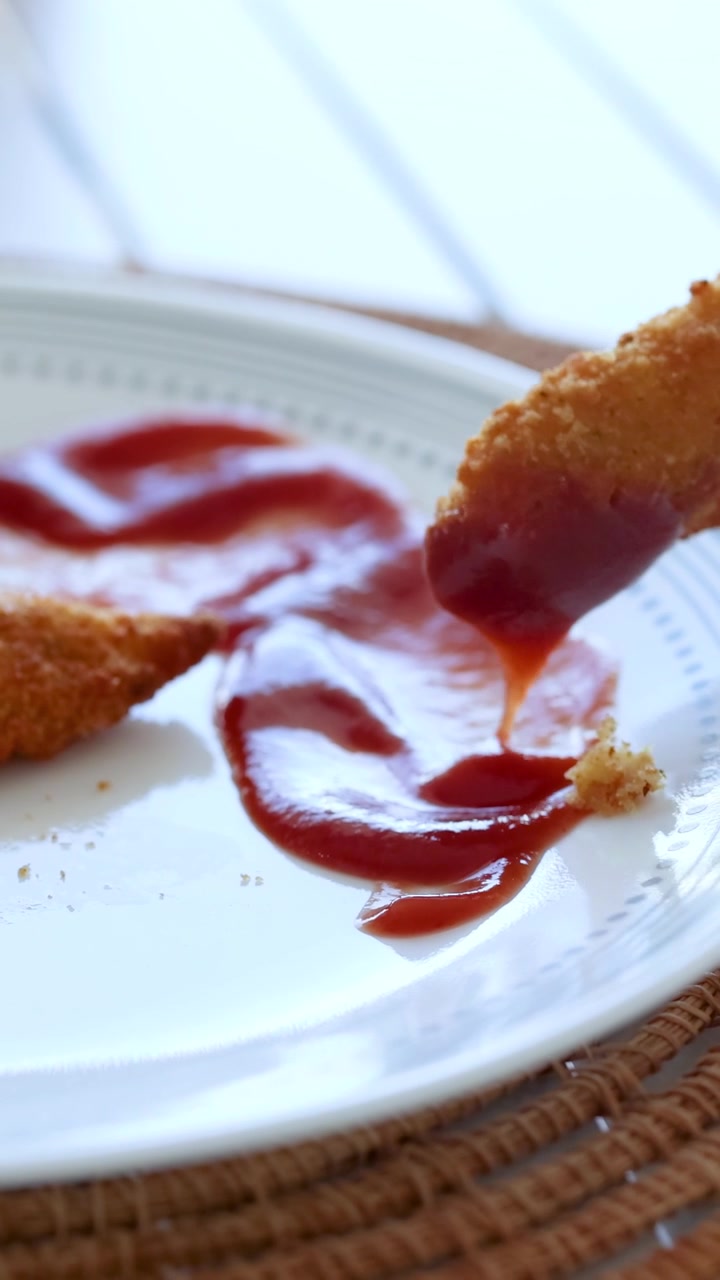 Una mano intinge pollo fritto nel ketchup su un piatto bianco, sotto una luminosa luce naturale.