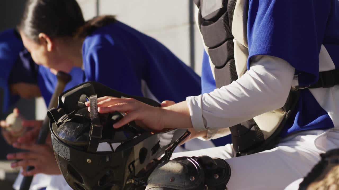 헬을 입고 팀과 함께 벤치에 앉아 있는 백인 여성 야구 선수