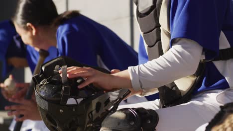 jugadora de béisbol caucásica sentada en el banco con el equipo, usando casco