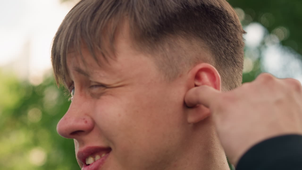 야외에서 귀를 잡아당기는 젊은 백인 남성, 십 대 사이의 장난, 억지웃음과 찡그린 표정, 손가락으로 귓불을 잡아당김, 잎이 무성한 공원 배경, 솔직한 반응