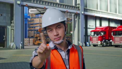 close up of asian male engineer with safety helmet disapproving with no index finger sign while standing , outside of logistics distributions warehouse