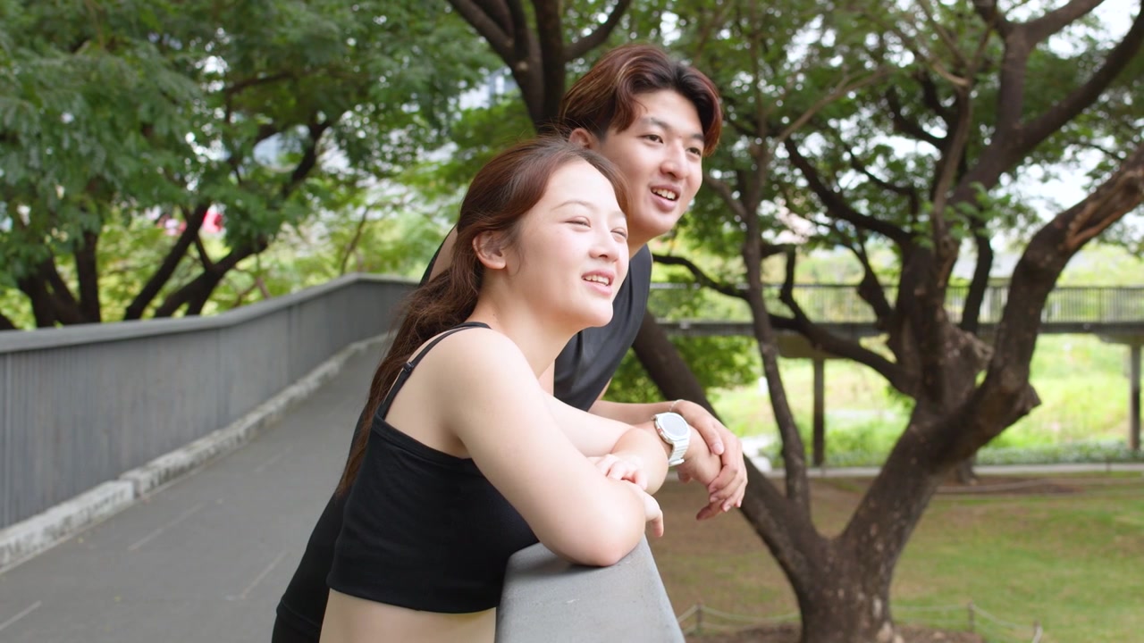 黒いスポーツウェアを着た若いアジア人カップルが、手すりに寄りかかり、外を指差しながら話している。