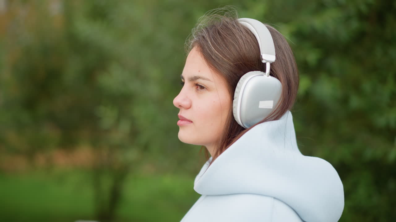 Close-up side view of young woman with headphones, listening to music outdoors in a garden with a blurred background of trees, perfect for videos on relaxation, music, or nature enjoyment