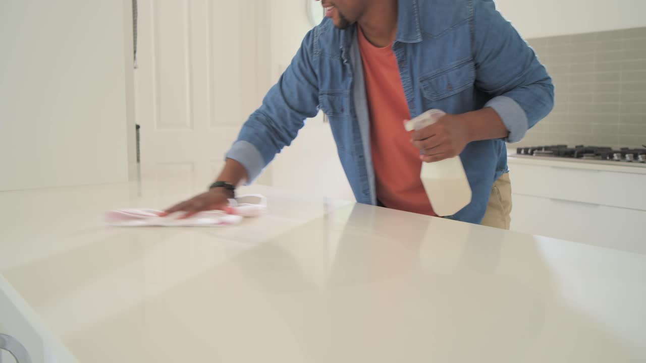 African American man approaching kitchen island spraying bottle wiping cloth cleaning, copy space