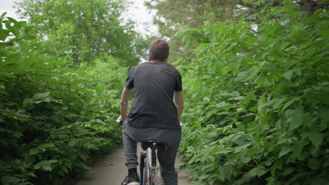 back view of young boy wearing black top and grey trouser cycling along a paved path with white road markings, surrounded by vibrant greenery that forms a natural tunnel effect