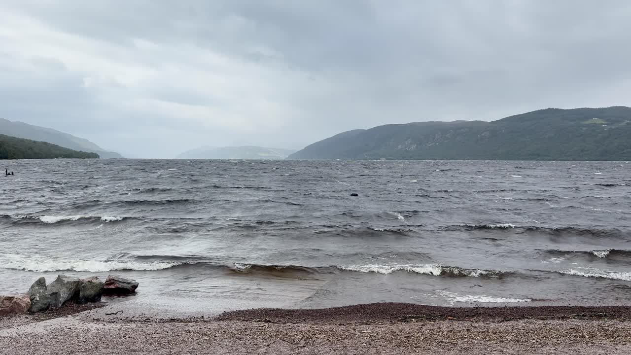 Choppy waves hit rocky Loch Ness shore under overcast skies, static wide shot, natural daylight