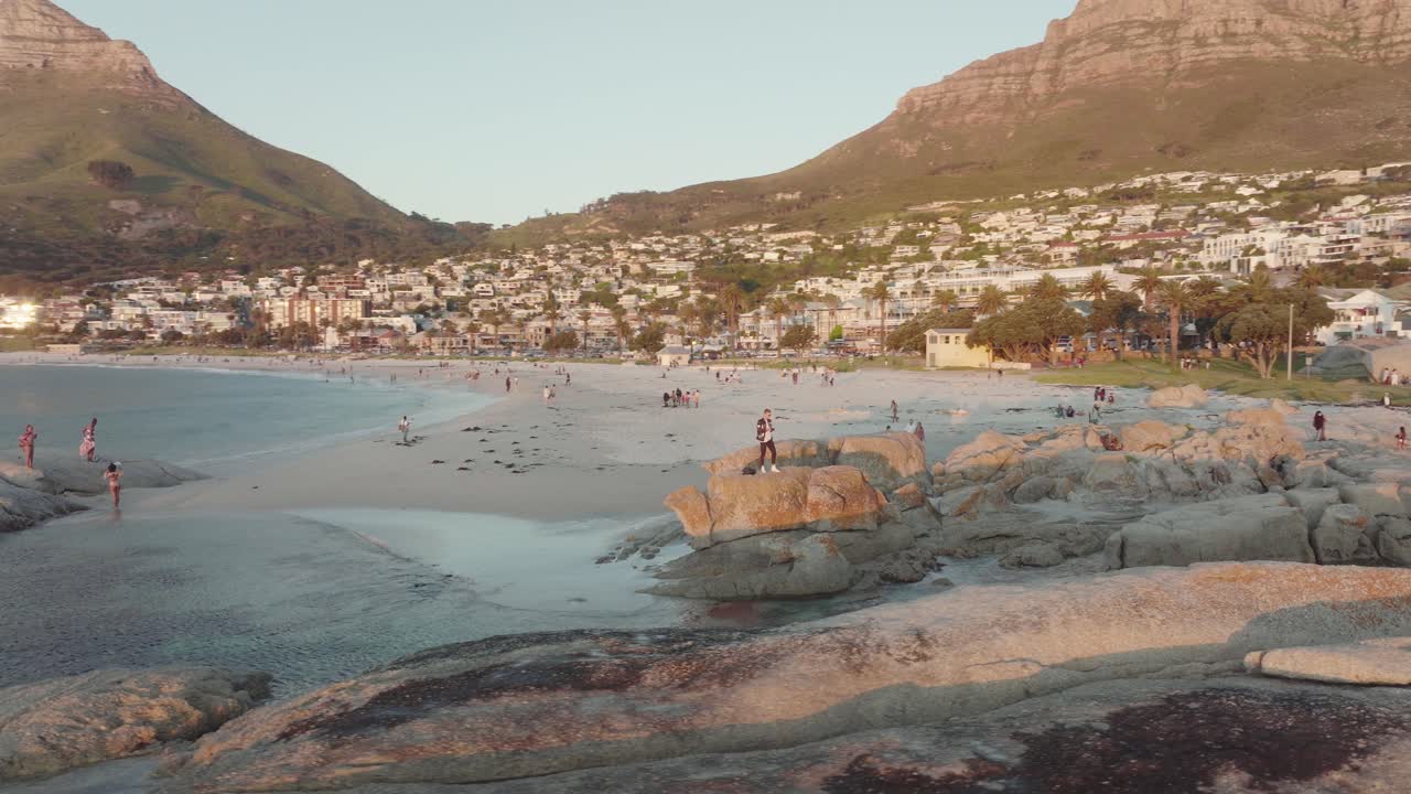 Drone flying around big rocks at the beach of Camps Bay in Cape Town, South Africa - in the background the Lion's Head Mountain rises illuminated by the sunset - Many houses on the hill