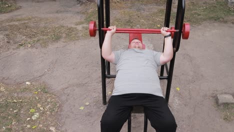 a persistent fat man lifts weights lying down on outdoor exercise machines a persistent fat man lifts weights lying down on outdoor exercise machines struggle with excess weight training man on the background of asphalt