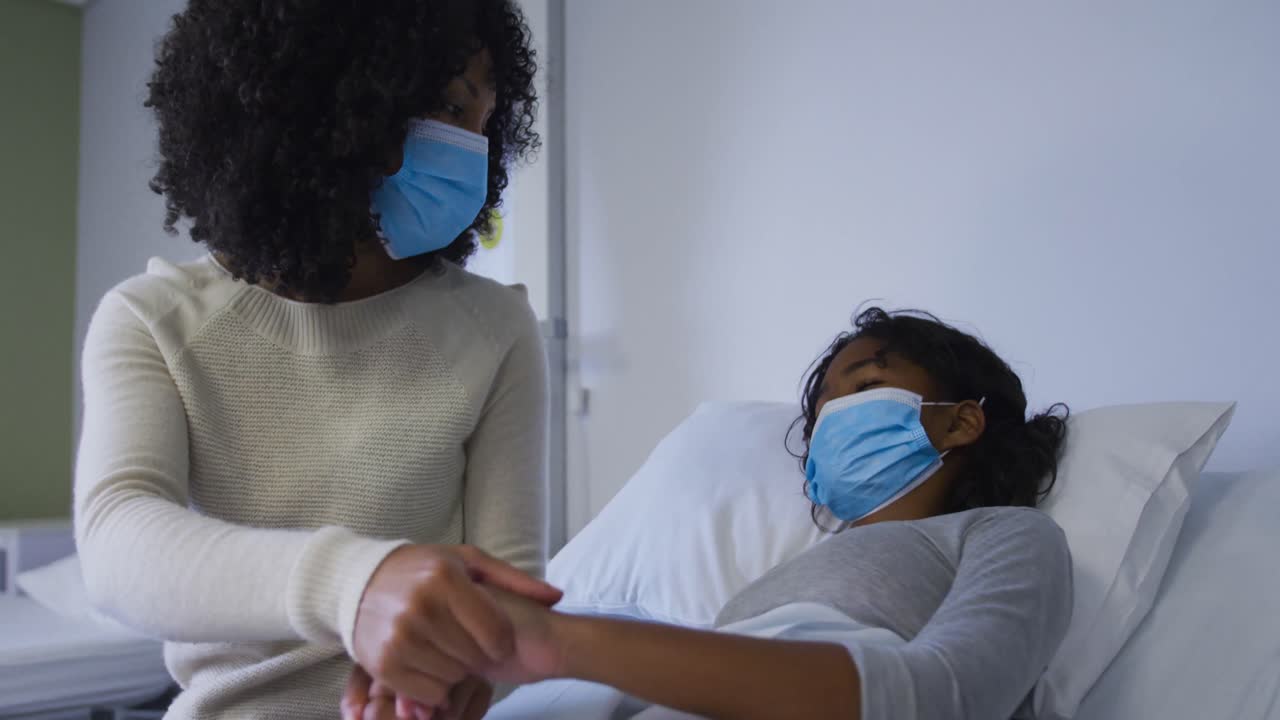 African american mother wearing face mask holding hands and comforting her daughter at hospital