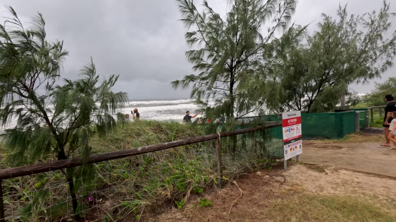 사람들은 구름이 가득한 하늘 아래 해안 경로를 따라 걸어가고, 사이클론의 위험성을 나타내는 경고 표지판이 있습니다.
