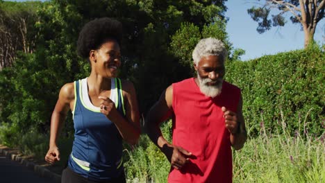 smiling african american senior couple exercising outdoors running in sunny green road