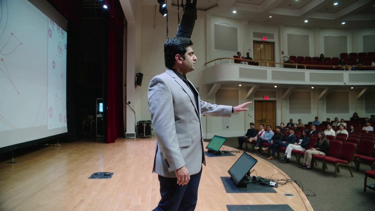 Starting beside left screen, presenter in gray blazer presenting, walking and gesturing to audience