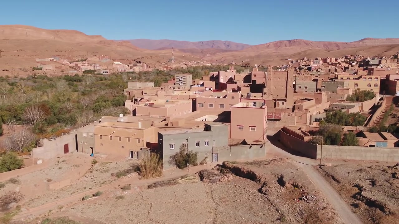 Veduta aerea cinematografica a bassa quota che segue le acque che scorrono del fiume Todgha, il quale serpeggia oltre lussureggianti alberi di oasi e un villaggio in terra battuta a Tinghir, Marocco