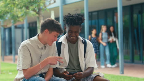two male high school or secondary students looking at social media on phone sitting outdoors