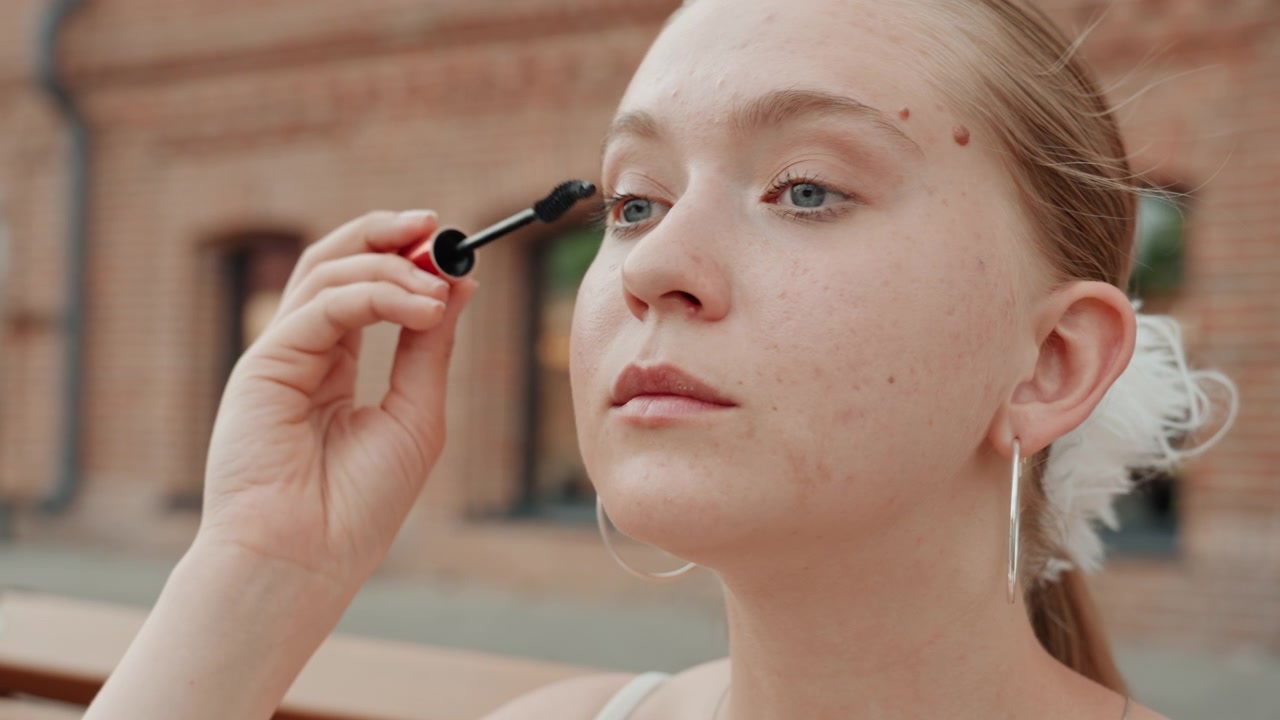 Young woman applies mascara outdoors in front of brick building. Woman holds red and black product focusing on her eye makeup with soft natural light illuminating her skin and jewelry