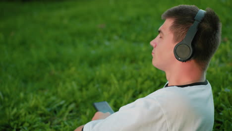 man lies on his side in grassy field enjoying music outdoors, resting on his hand with headphones, looking relaxed and content in a peaceful natural environment