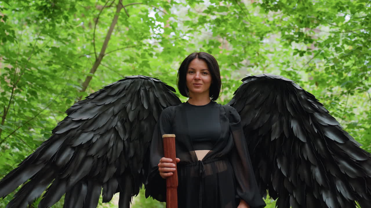 Donna elegante che indossa un costume di piume nere mentre manda un bacio leggero con un sorriso accennato, tenendo in mano un bastone di legno in una lussureggiante foresta verde, circondata dalla luce del sole e dal fogliame, creando un’atmosfera di fantasia